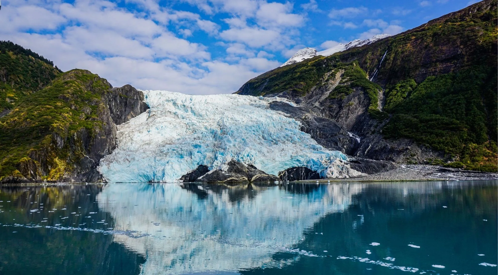 A tidewater glacier's blue calving face meeting the turquoise water of a Prince William Sound fjord, with brash ice drifting in the foreground