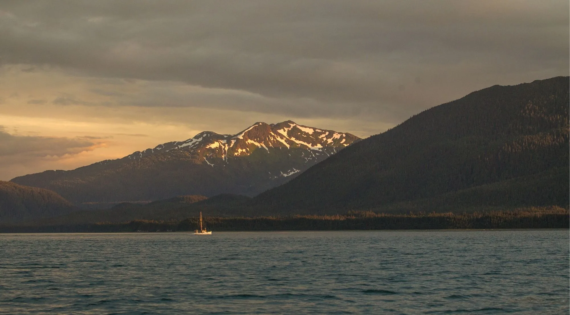 A sailboat silhouetted on Prince William Sound at sunset, with golden light on the ridgeline and dark mountains behind