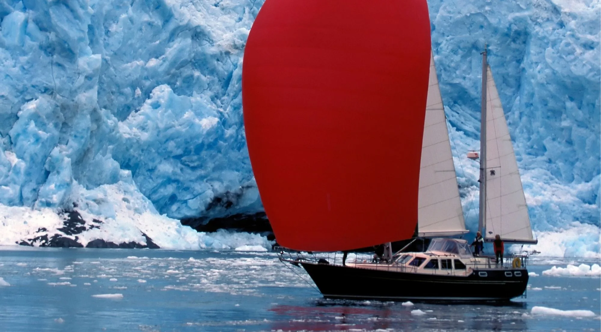 A sailing yacht with a red spinnaker passing close to a towering blue glacier face streaked with crevasses