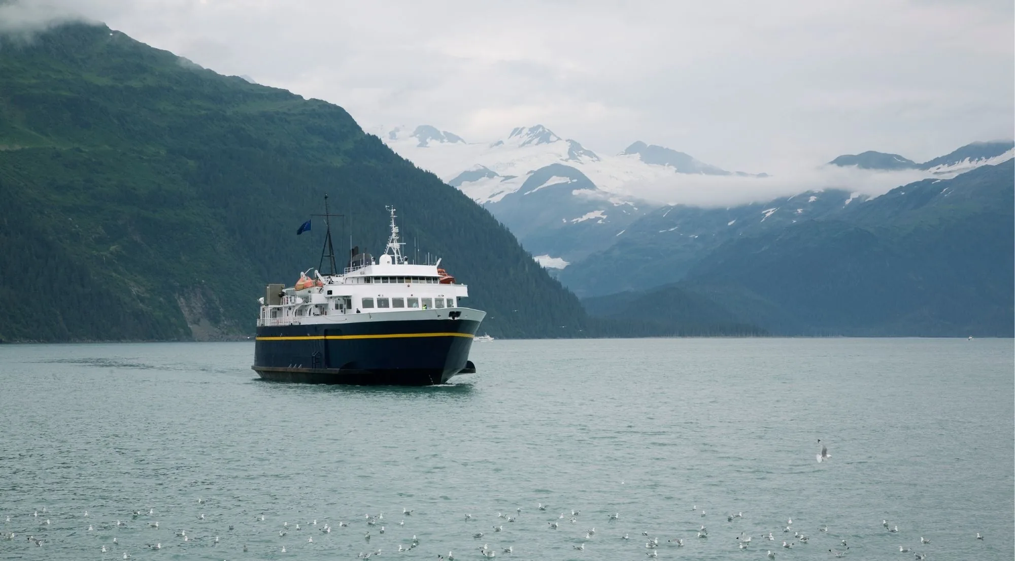 A blue and white tour vessel crossing a glassy fjord with snow-covered peaks and a glacier valley behind it