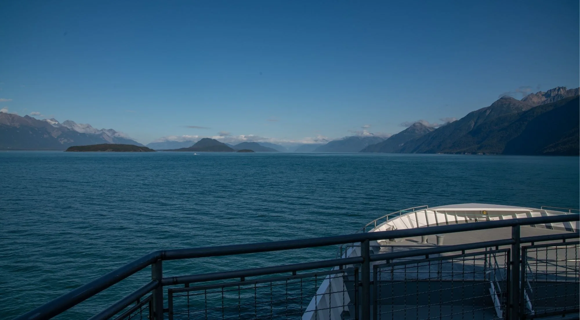 The view from a tour boat's stern railing across deep blue water toward distant snow-capped mountains