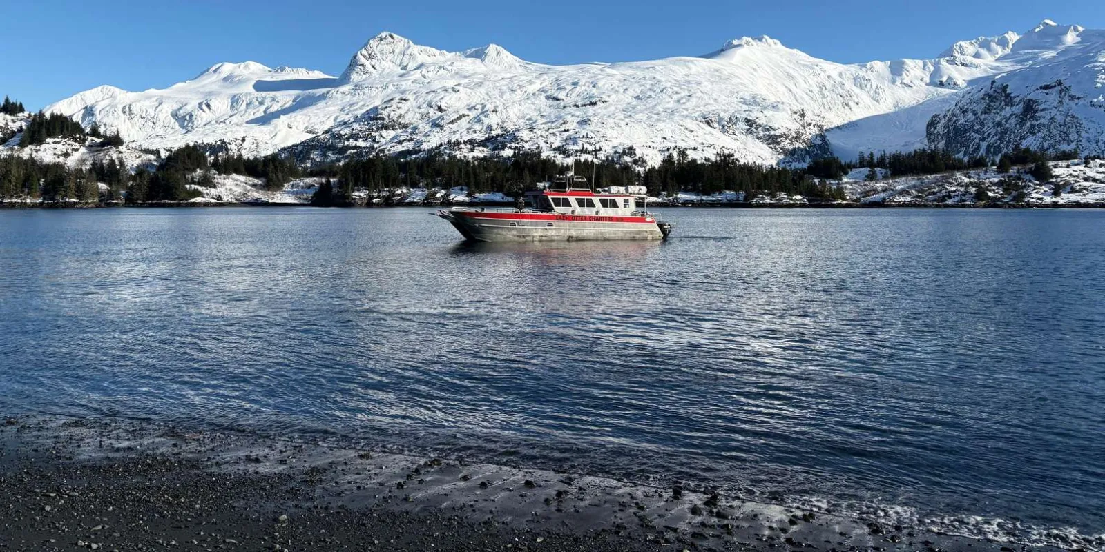The small Lazy Otter Charters vessel anchored off a dark gravel beach below snow-covered peaks in Blackstone Bay