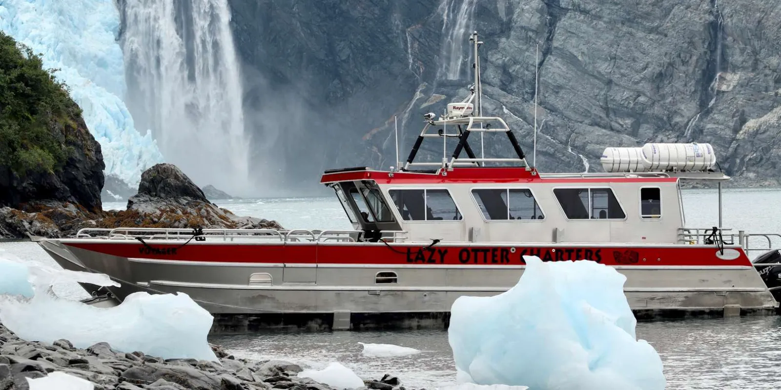 The Lazy Otter Charters boat pulled in close to a remote shoreline below a tall waterfall, with glacier ice grounded on the beach