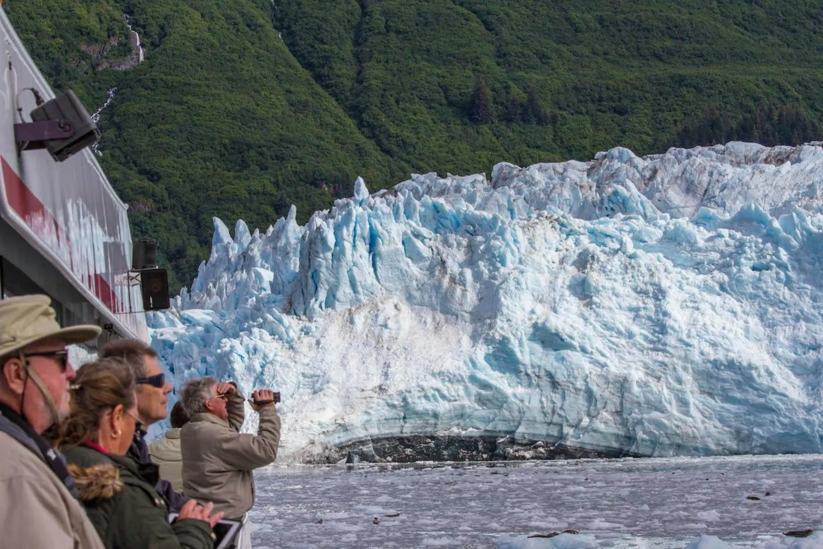 Passengers watching from the deck rail as the boat sits close to the jagged blue ice front of Meares Glacier