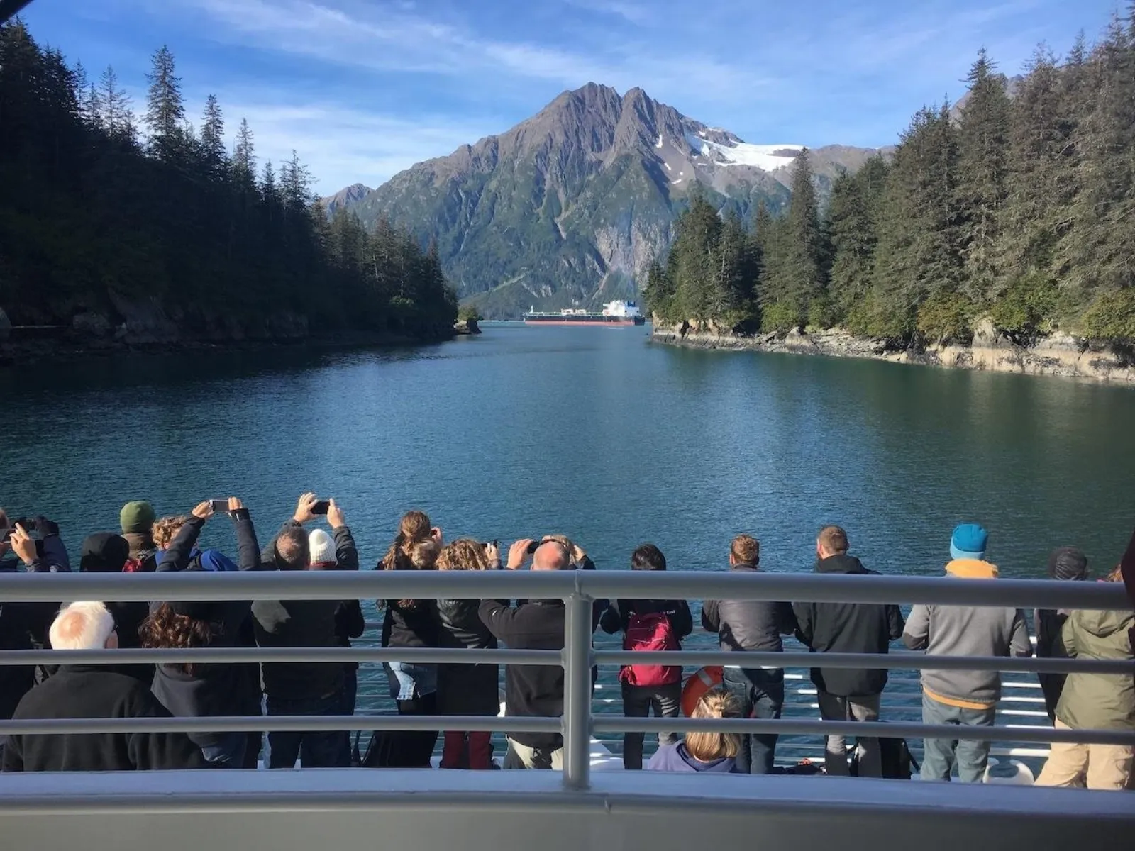 Passengers lining the deck rail to photograph a forested inlet with a snow-streaked peak at its head