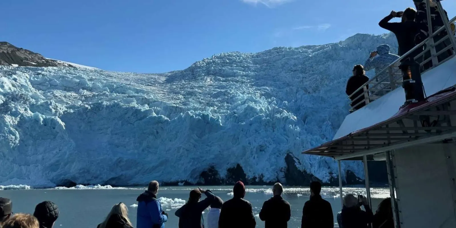 Passengers silhouetted along the boat's upper deck photographing a wide glacier front across the water
