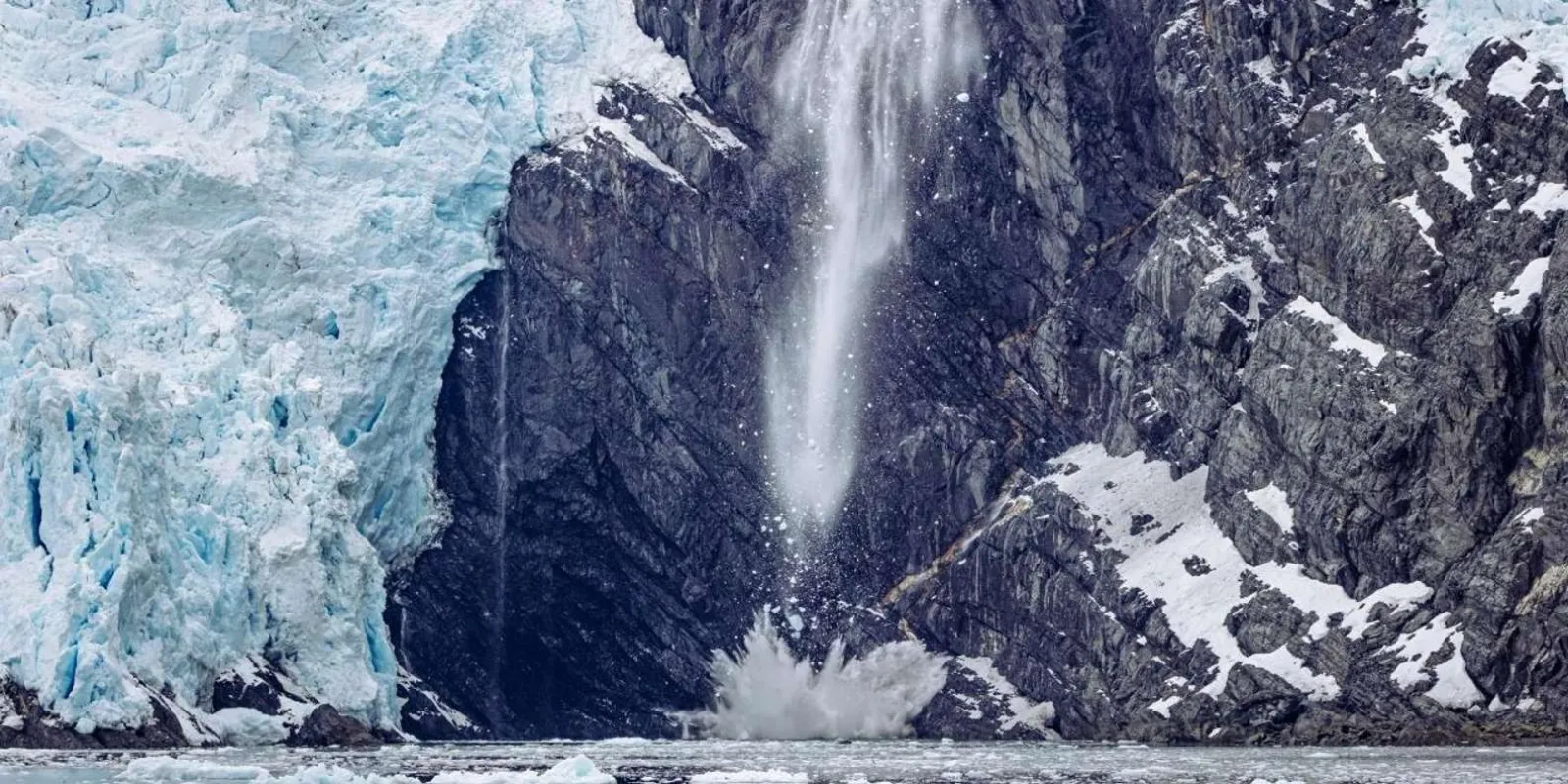 A tall waterfall plunging down a dark rock face beside the blue ice of a glacier at the waterline