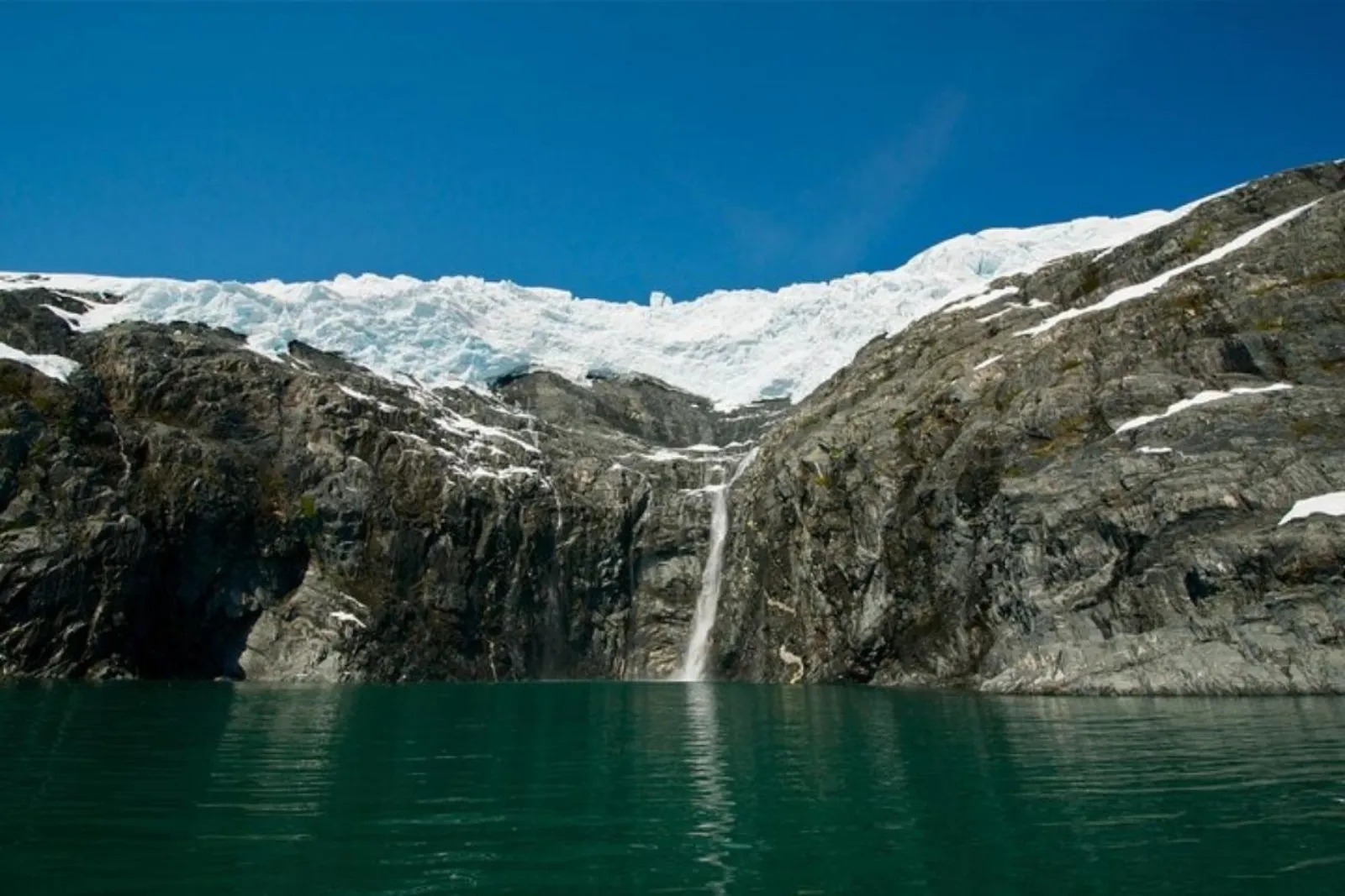 A tidewater glacier spilling over a dark rock headland into the green water of a Prince William Sound fjord under a clear blue sky