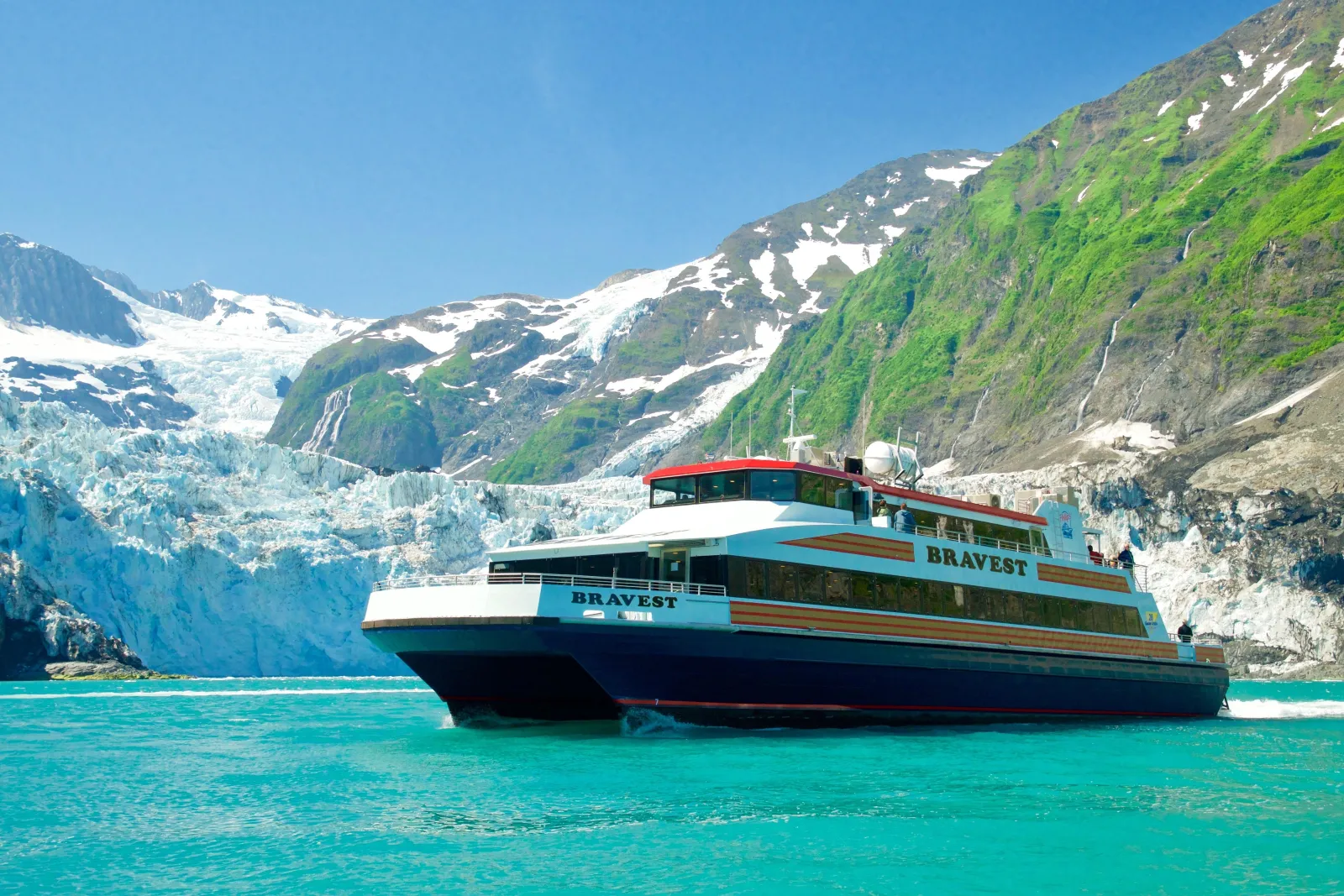 The catamaran Bravest holding position on turquoise water below a broad glacier and green mountain slopes streaked with waterfalls